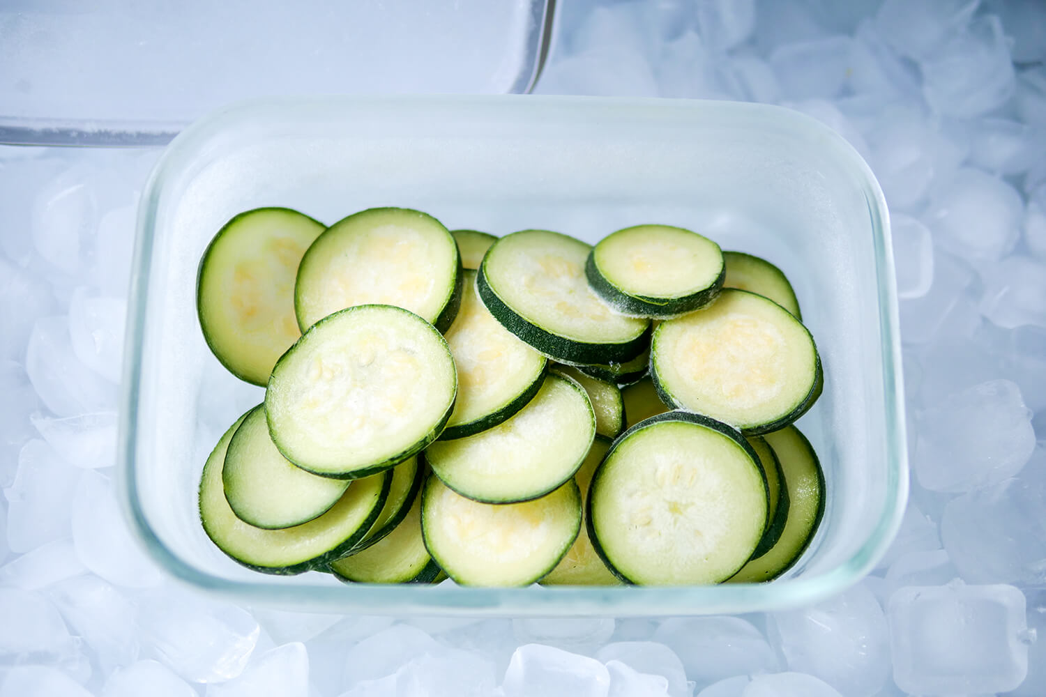 In Scheiben geschnittene, gefrorene Zucchini in einer Glasschüssel, umgeben von Eiswürfeln im Gefrierfach.