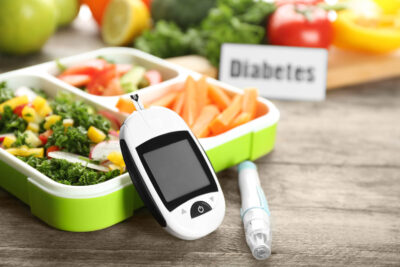 Digitales Blutzuckermessgerät, Lanzettenstift und Lunchbox mit Gemüse auf dem Tisch.