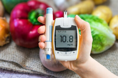 Diabetische Ernährung: Hand hält Glukometer mit Gemüse im Hintergrund.