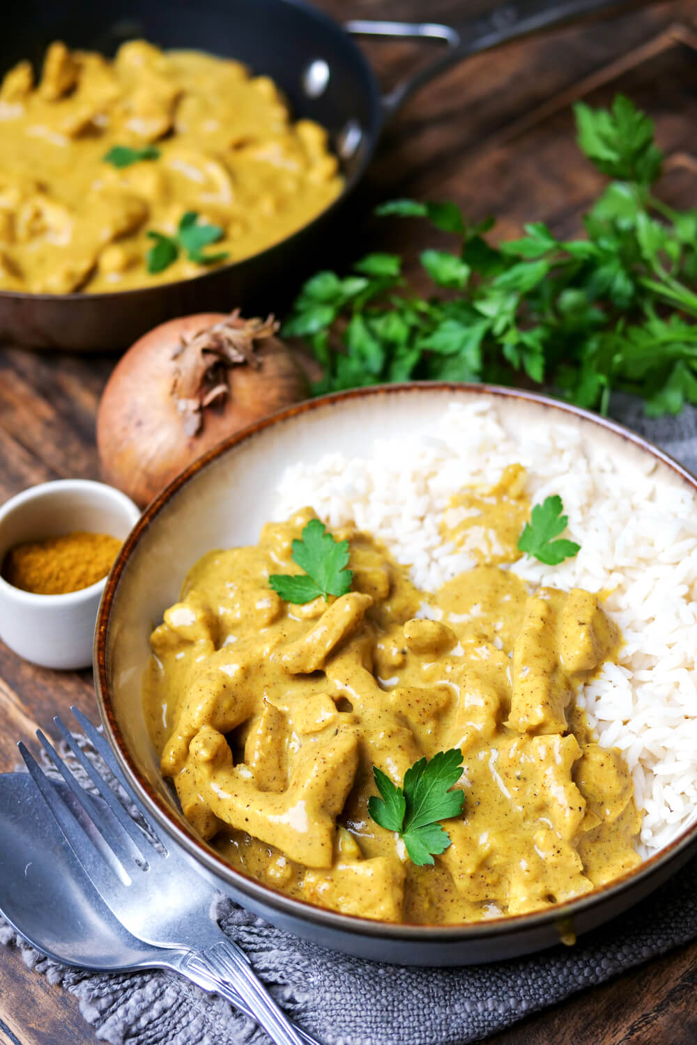 Curry-Geschnetzeltes mit Reis und Petersilie auf einem Teller angerichtet, daneben Besteck angelegt.